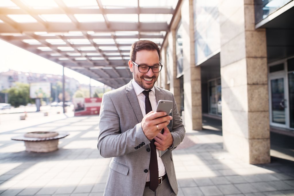 Ein lächelnder Unternehmensberater im grauen Anzug und mit Brille hält sein Smartphone und ein Tablet. Er steht vor einem modernen Bürogebäude und ist mobil vernetzt.